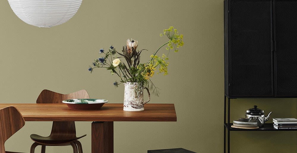 Spisebord i mørkt tre mot olivengrønn vegg, hvit papirlampe, mugge med villblomster.