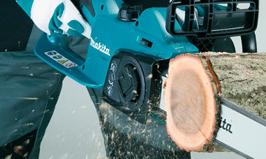 Hender med hvite og grønne hansker holder turkis Makita-kjedesag som sager i tre, flis spruter