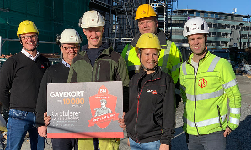 Seks byggarbeidere med gule og hvite hjelmer holder rødt og grått gavekort-skilt foran en byggeplass.