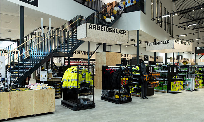 Interiør av lys byggevarebutikk med gule arbeidsjakker under ‘Arbeidsklær’-skilt og verktøyhyller