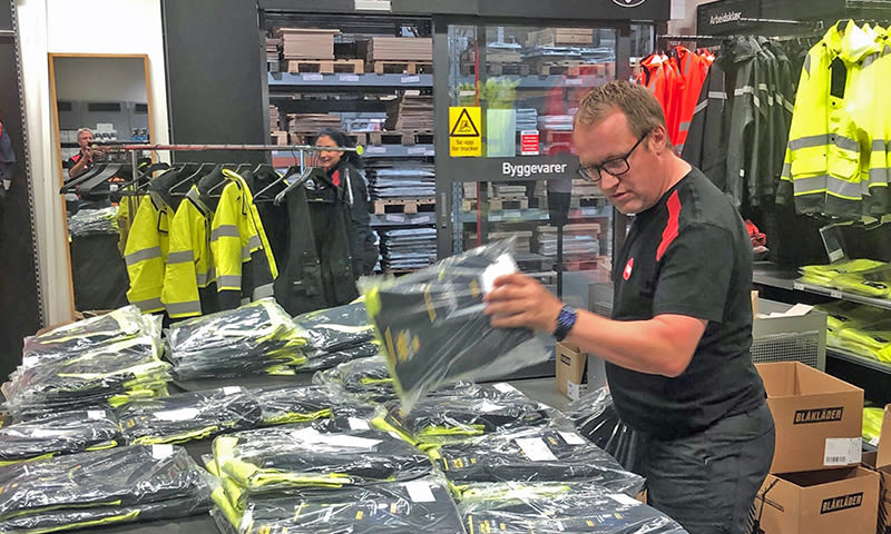 Ansatt stabler plastinnpakkede gule og svarte arbeidsklær på bord i butikk.
