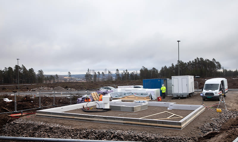 Lav byggeplass med husfundament på grus, blå container, hvit varebil og skog under grå himmel.