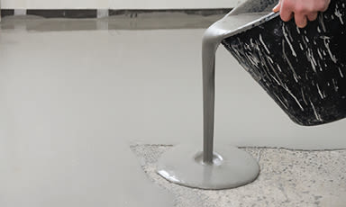 Hånd heller grå selvnivellerende masse fra svart bøtte på betonggulv.