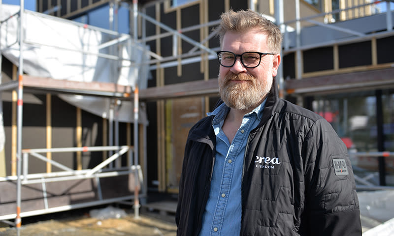 Mann i svart jakke på byggeplass med stillas og uferdig fasade.