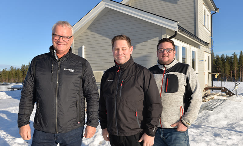 Tre menn i vinterjakker foran nybygd grå trevilla, snø og furuskog under blå himmel