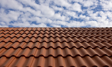 Rødbrunt bølget tak under blå himmel med spredte hvite skyer.