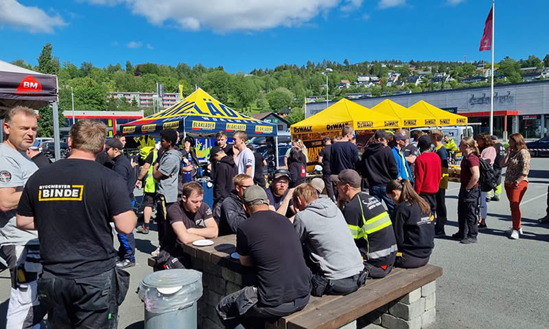 Folk i arbeidsklær ved gule Dewalt-telt på solfylt parkeringsplass med grønne åser bak