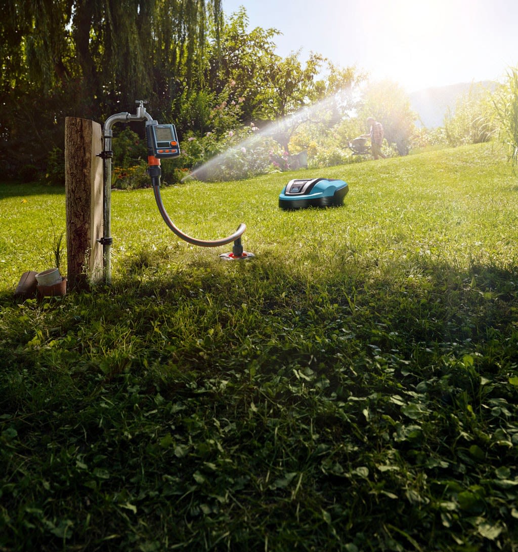 Turkis robotgressklipper på solbelyst plen, vannstråle fra hageslange ved kran.