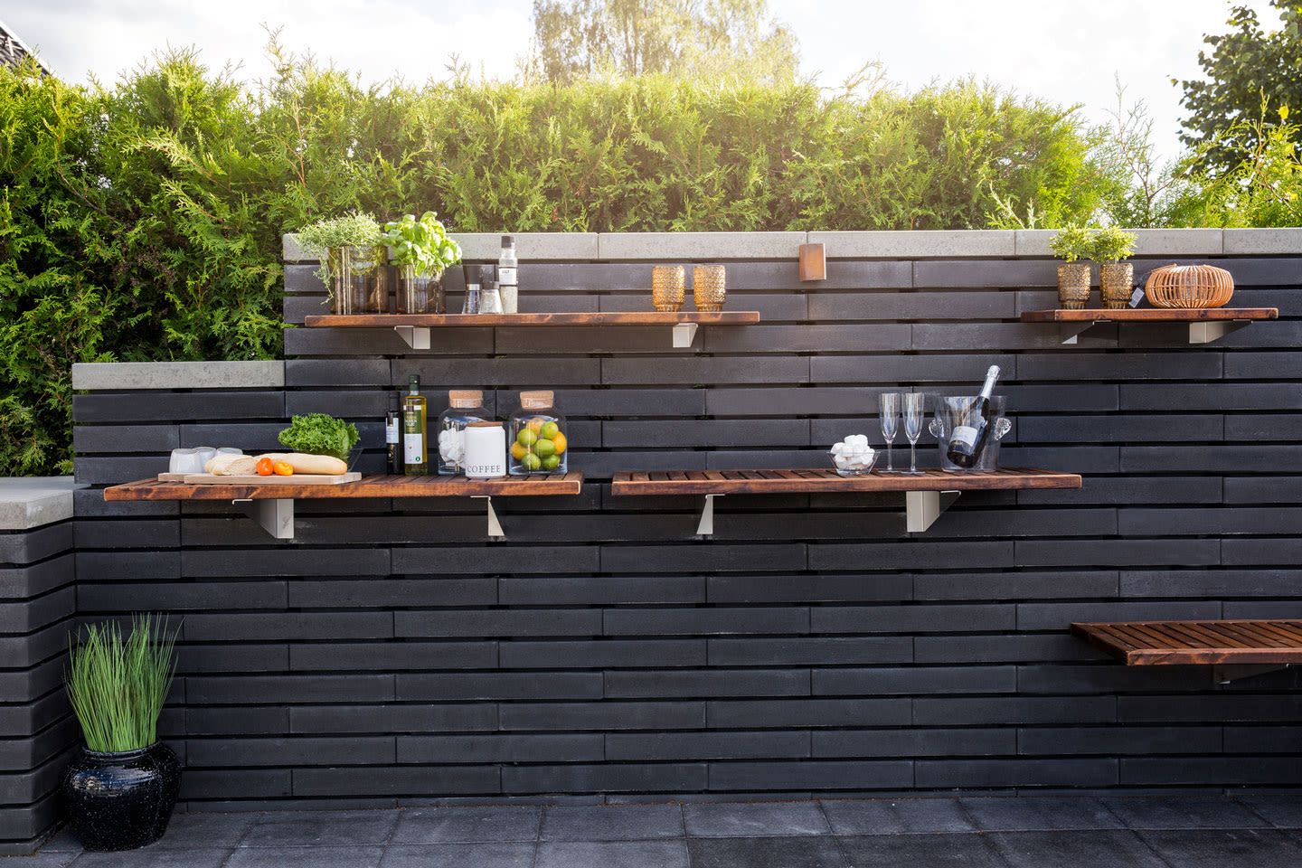 Mørk murvegg med flytende trehyller pyntet med urter, frukt, kjøkkenutstyr og champagneglass.