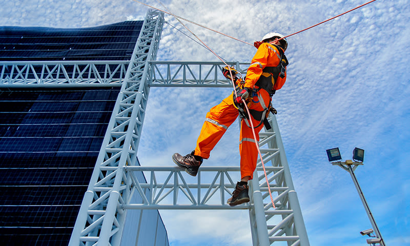 Arbeider i oransje vernedrakt klatrer i stålramme ved store solcellepaneler under blå himmel