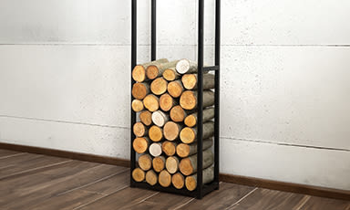 Svart metallstativ med stablet vedkubber foran hvit vegg og tregulv.