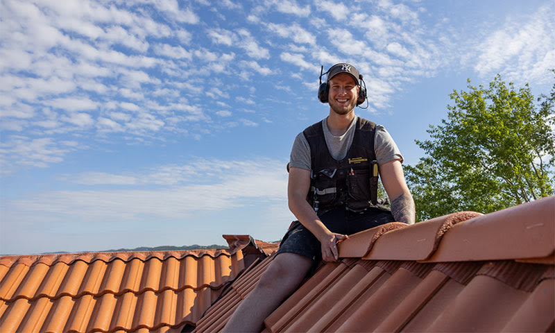 Takarbeider med vernevest og hørselsvern legger rød takstein under blå himmel med lette skyer.