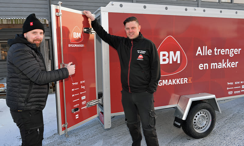 To ansatte i svarte jakker ved rød Byggmakker-tilhenger med teksten «Alle trenger en makker» på snø.