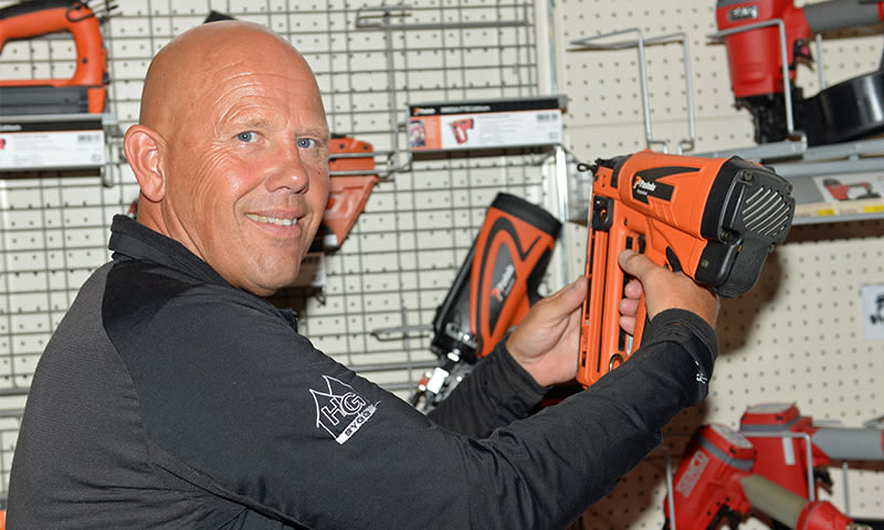 Person i svart jakke holder oransje spikerpistol foran hylle med verktøy