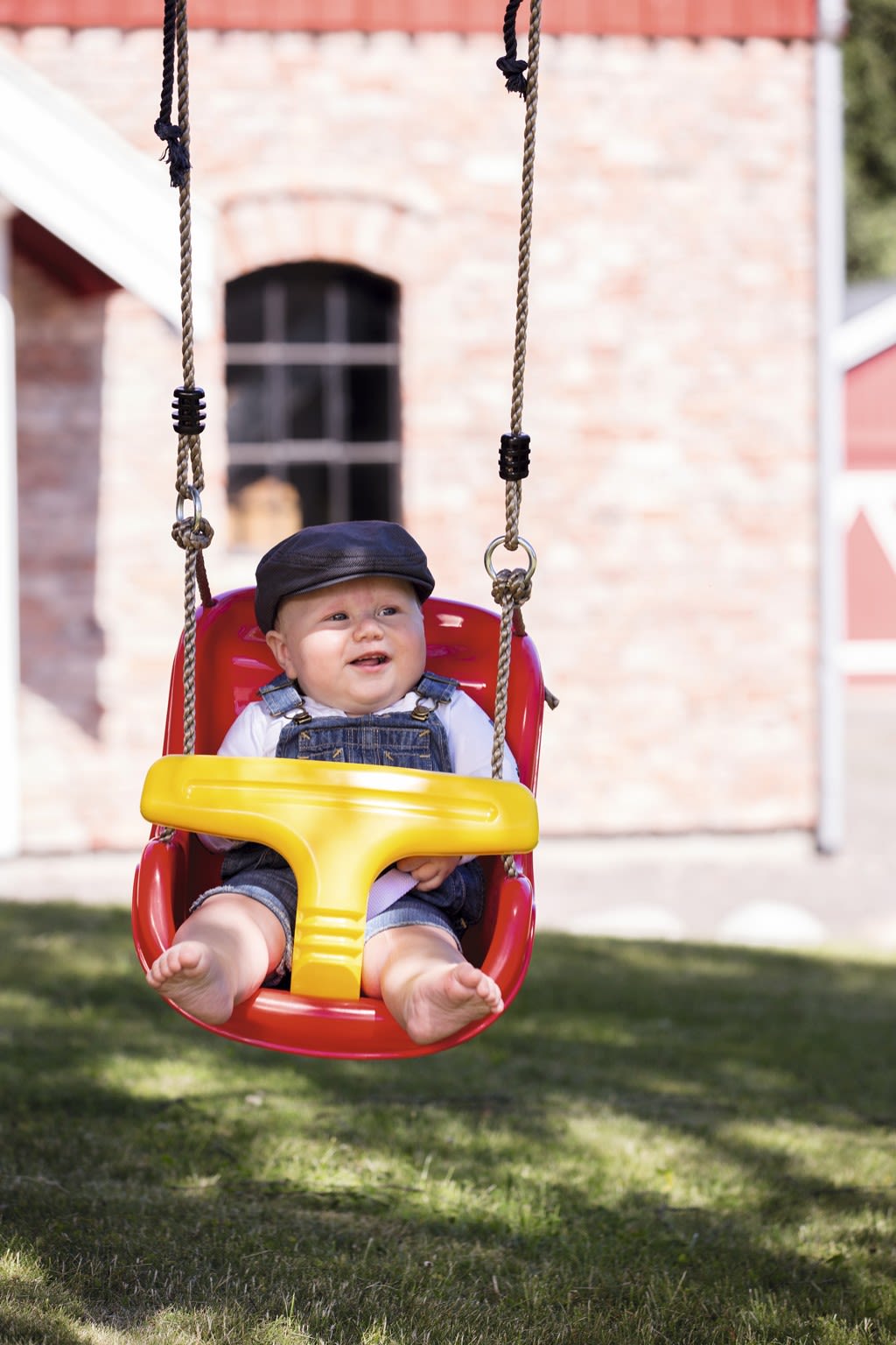 Småbarn i denimkjeledress og mørk lue i rød-gul plasthuske foran murvegg i hage.