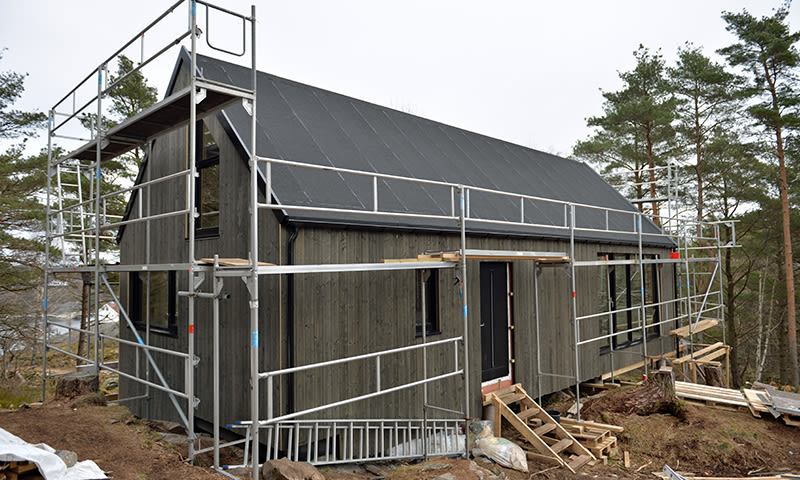 Mørk trehus under oppføring med stillas, svart tak og furutrær i bakgrunnen.