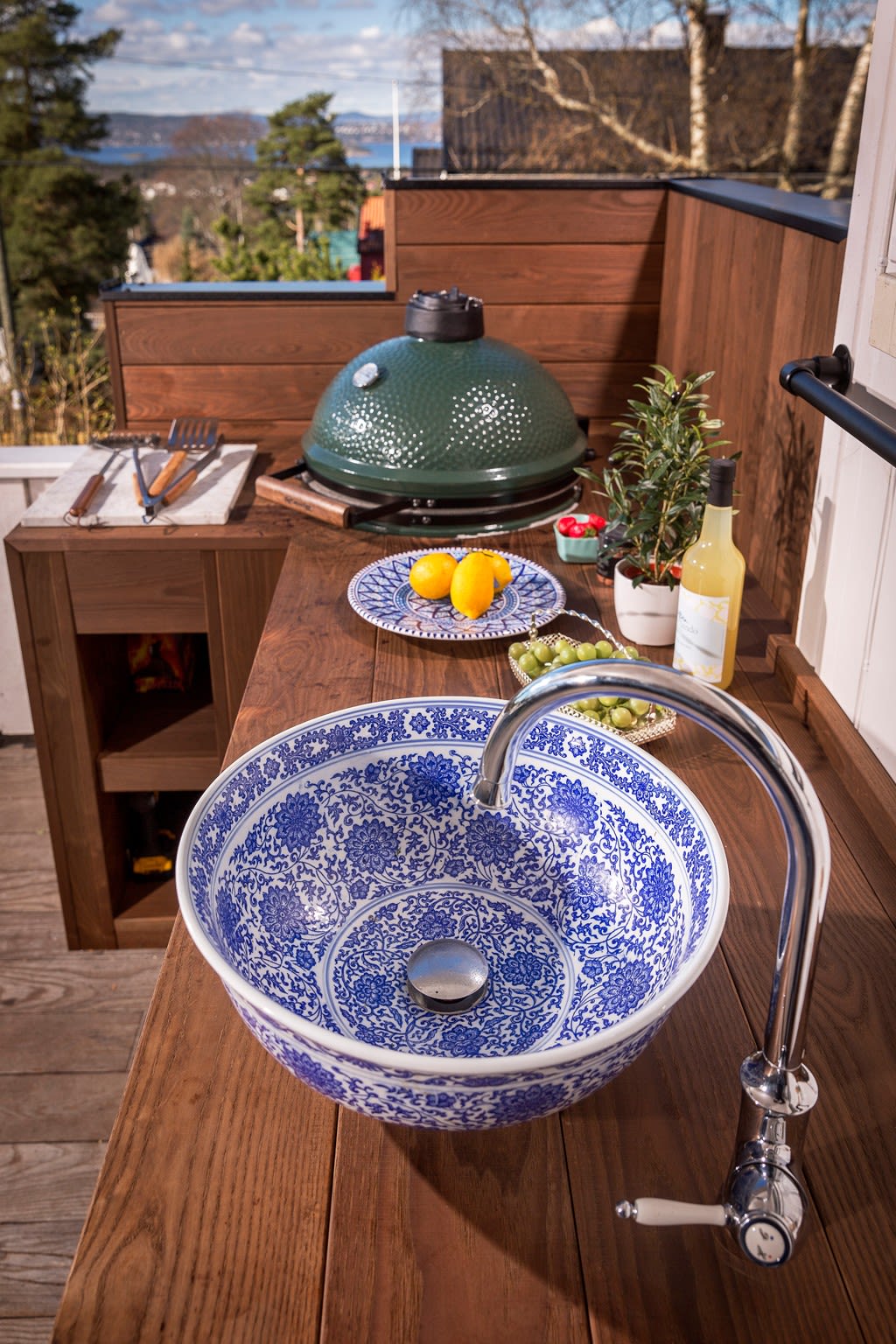 Utendørs benk med blå-hvitt porselensvask, kromkran, grønn grill, sitroner, druer og flaske.