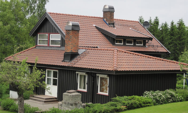 Svart trehus i to etasjer med rødt tegltak, to skorsteiner og grønn hage i skogkanten.