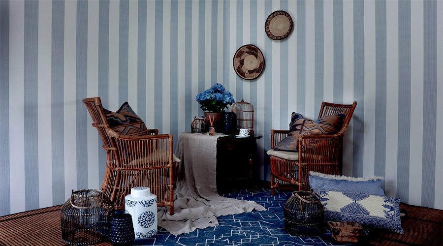 To rottingsstoler med puter, blått teppe, grå-hvitt stripet tapet og bord med blå hortensia.