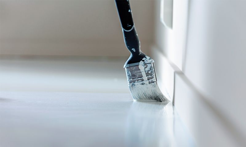 Pensel med svart håndtak maler hvit gulvlist langs vegg