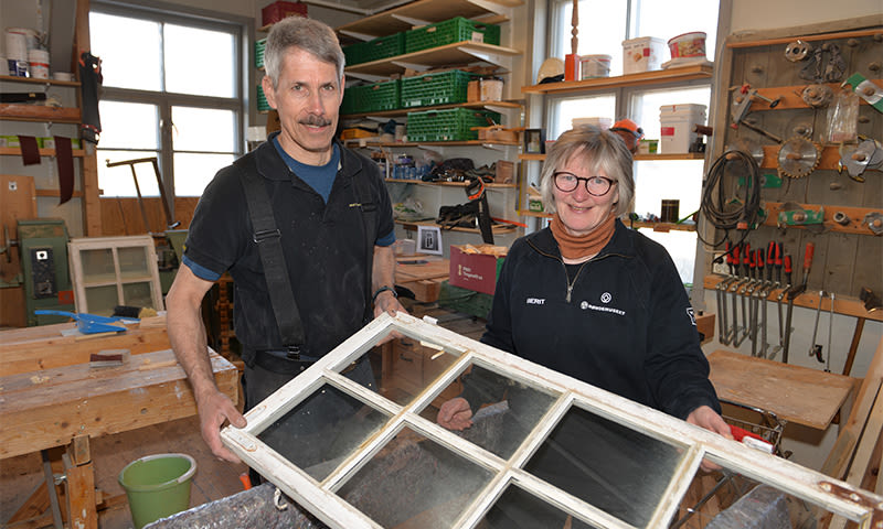 To personer i lyst treverksted med grønne bokser og verktøy holder en gammel vindusramme