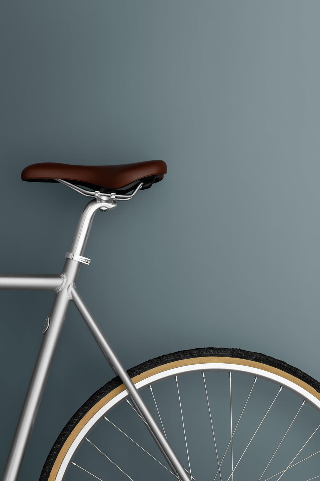 Sykkelbakhjul og sete mot blågrå bakgrunn, sølvgrå ramme, brunt skinnsete og svart/beige dekk