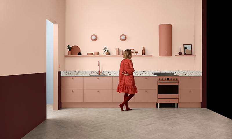 Kvinne i rød kjole i rosa minimalistisk kjøkken med terrazzo benkeplate og burgunderdetaljer.