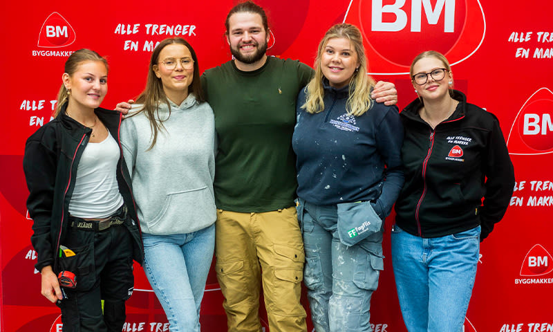 Fem Byggmakker-ansatte i arbeidstøy foran rød vegg med hvite BM-logoer.