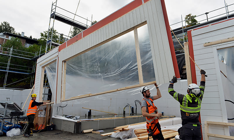 Tre arbeidere løfter en prefabrikkert hvit/rød vegg med stor vindusåpning på byggeplass.