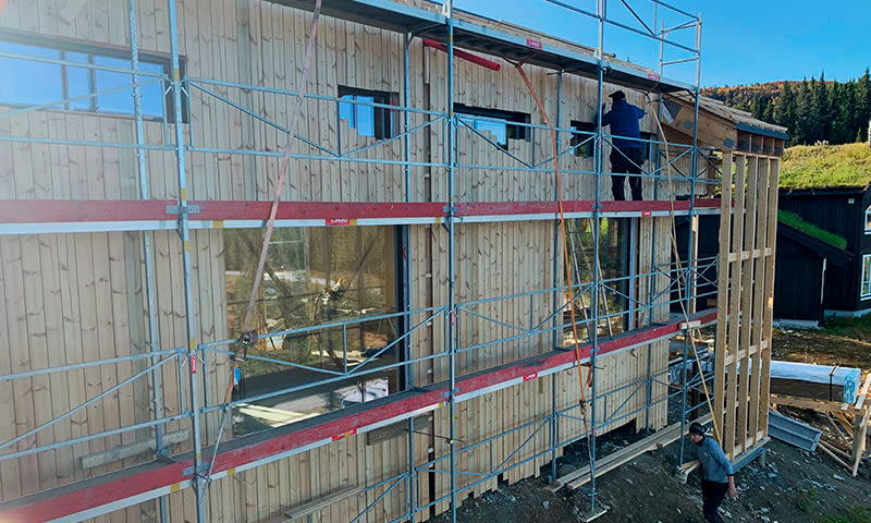 Arbeidere på stillas mot lyspanelert trehus under bygging mot blå himmel og skog.