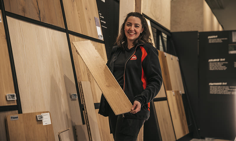 Ansatt i svart/rød jakke holder lys eikegulvplanke foran vegg med trepanelprøver.