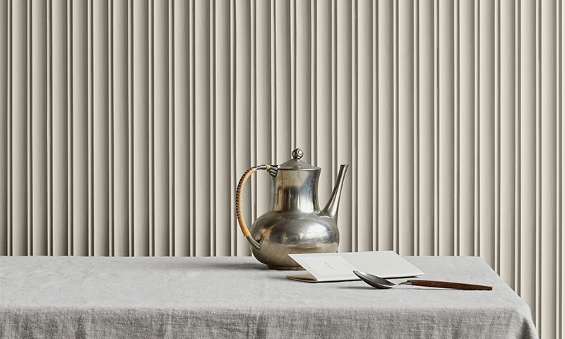 Sølv tekanne med flettet håndtak på grå duk, ved åpen bok og treskje foran beige plissert forheng.