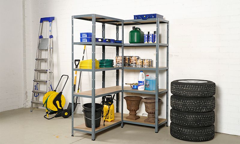 Grå hylle med hageredskaper, gul slange, grønne kanner, rengjøringsmidler og dekk foran hvit vegg