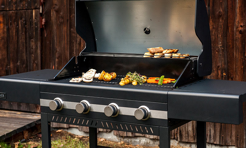 Åpen svart gassgrill mot tømmervegg med grillede grønnsaker, poteter, brød og urter.