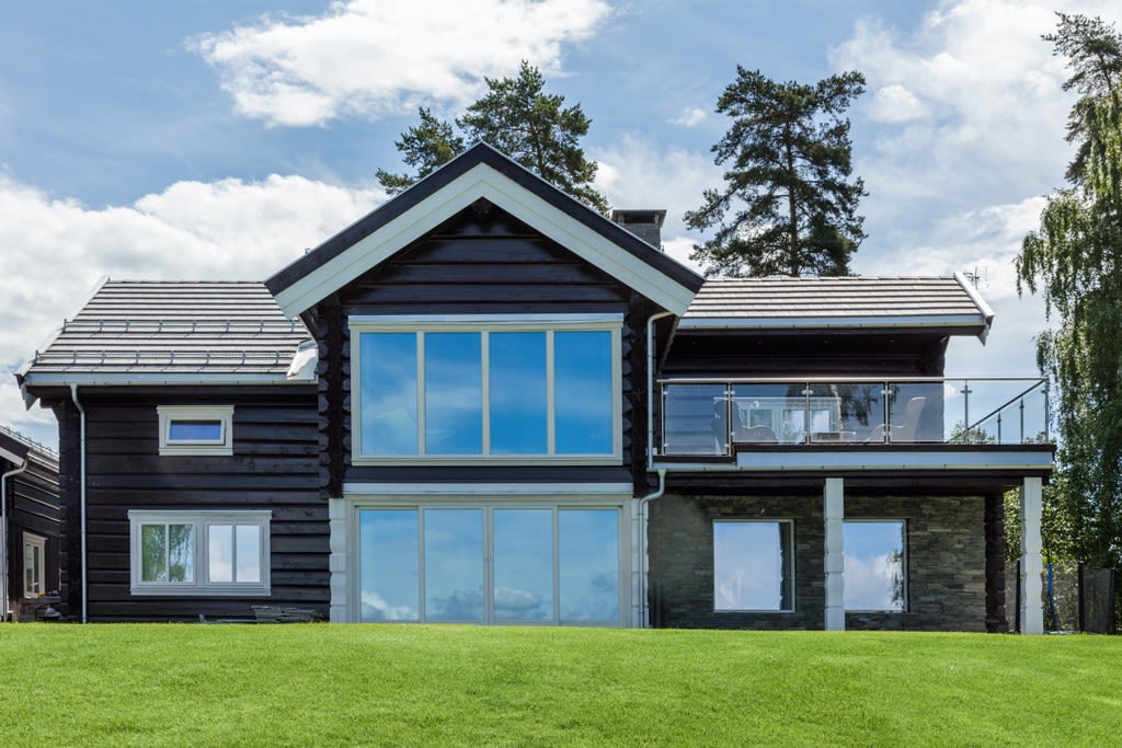 Moderne mørk trevilla med store vinduer, glassbalkong, grønt gress og blå himmel