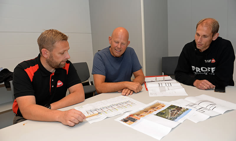 Tre menn i svart-rød, blå og svart genser ved hvitt bord studerer fargerike byggeplaner.