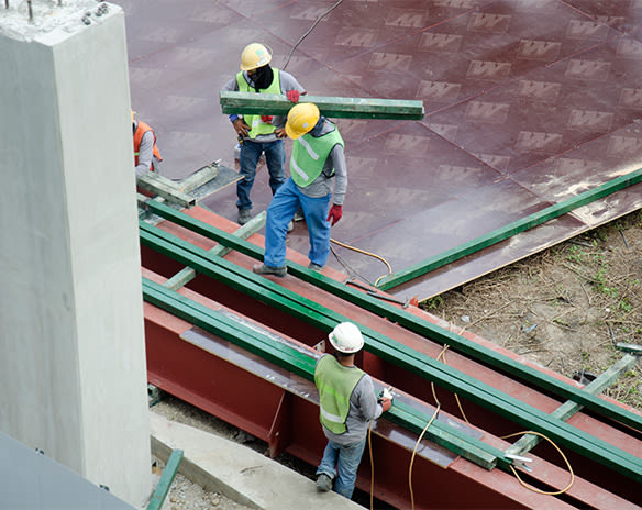 Tre arbeidere i gule hjelmer og grønne vester løfter stålbjelker på rødt rammeverk.