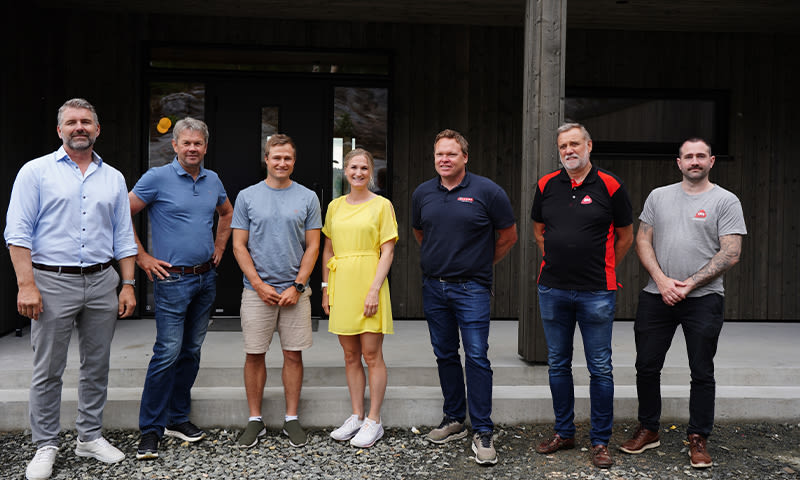 Sju personer i ulike fritidsklær står på grus foran moderne mørk trebygning.