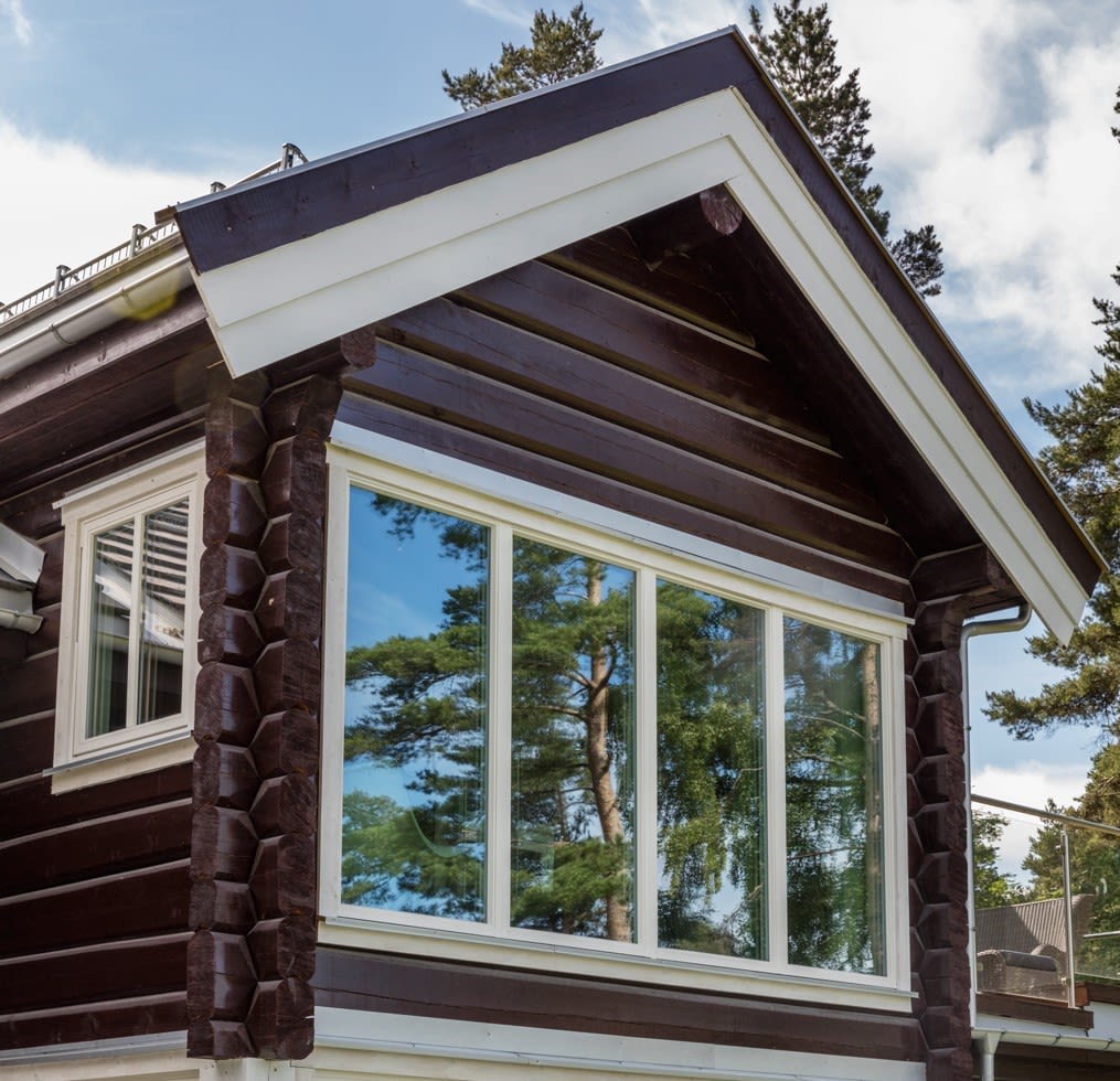 Brun tømmerhytte med hvite kanter og stort vindu som speiler grønne furutrær under blå himmel