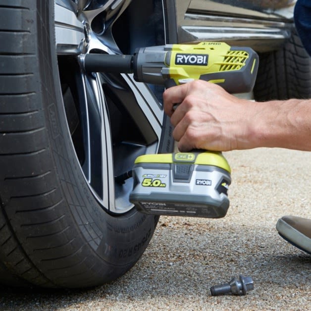 Hånd bruker gul-svart Ryobi muttertrekker på bilhjul på gruslagt underlag