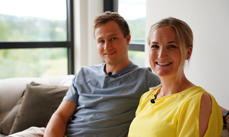 To personer i sofa foran vindu mot grønt landskap, en i gul topp og en i grå t-skjorte.