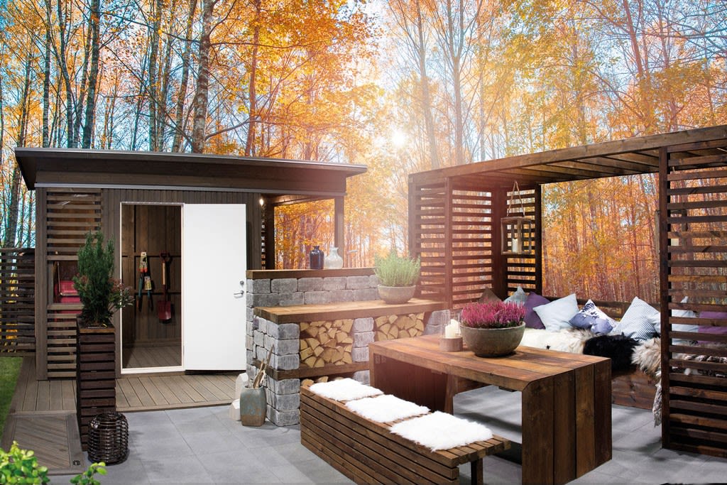 Uteplass med pergola i mørkt tre, steinbar, vedstabel og lyng i oransje høstskog