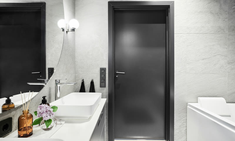 Moderne baderom med hvit vask, rundt speil, mørk grå dør, brun flaske og lilla blomster.