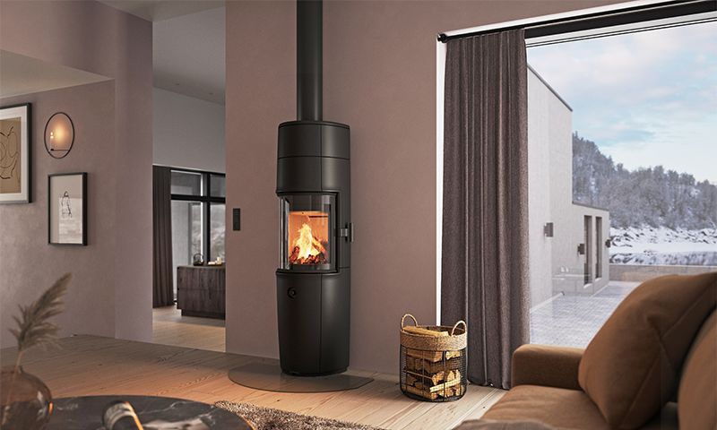 Koselig stue med svart sylinderovn, flammer, vedkurv og stort vindu mot snølandskap.
