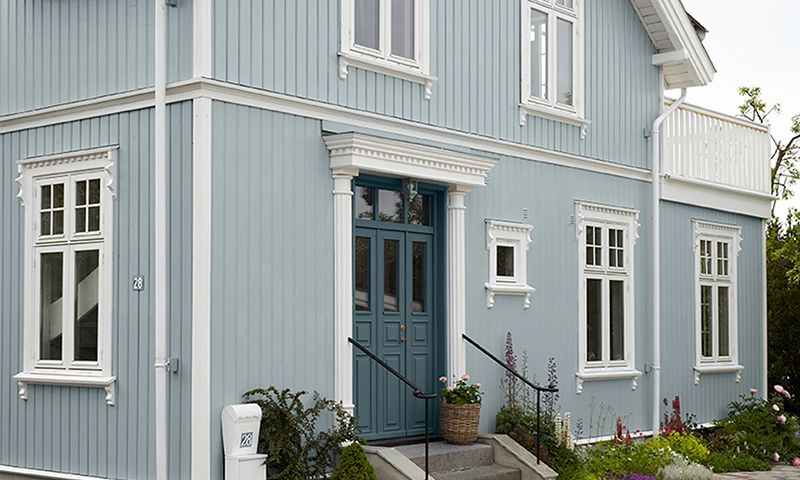 Lyseblå toetasjes trehus med hvite karmer, turkis dobbeltdør og blomster ved trapp