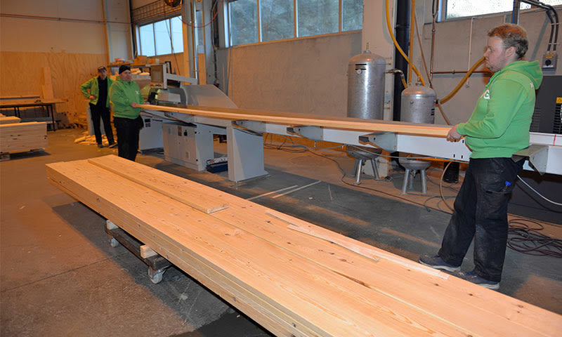 Tre arbeidere i grønn hettegenser fører lyse treplanker gjennom industriell sagmaskin i verksted