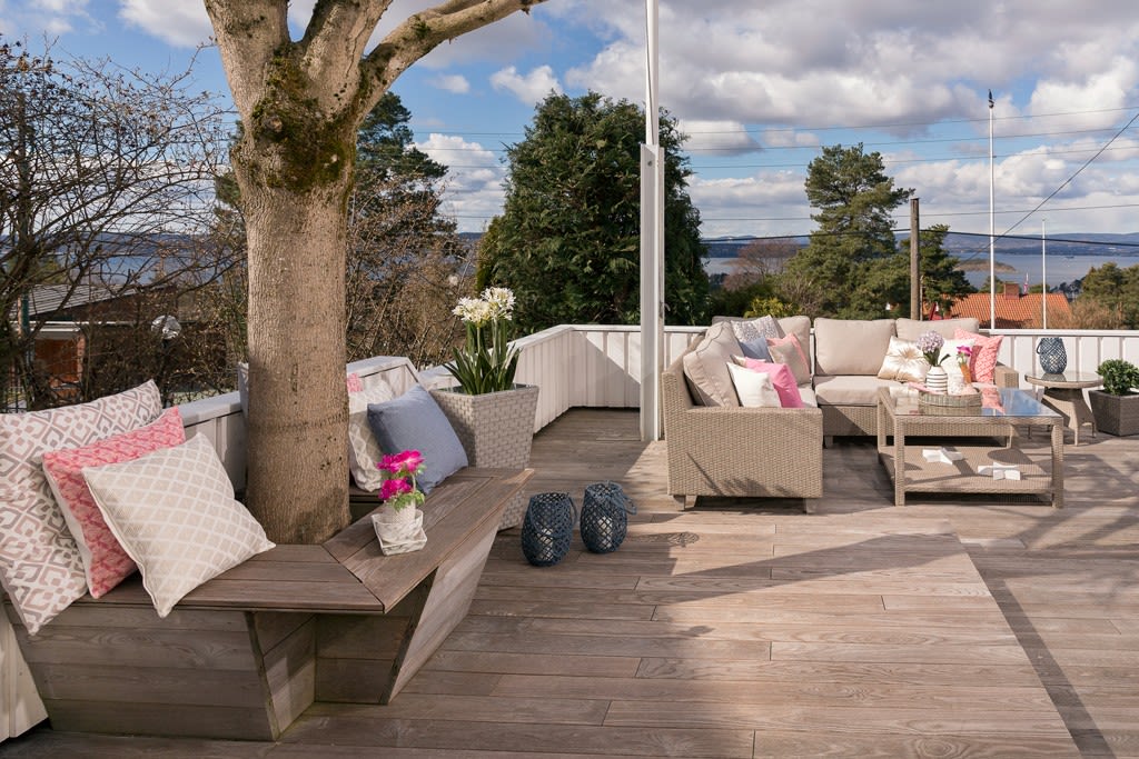 Solrik terrasse med tre i benk, rottingmøbler og rosa, hvite puter mot sjøutsikt.
