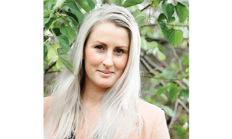 Person med langt sølvhvitt hår iført lys koralljakke foran grønt løvverk