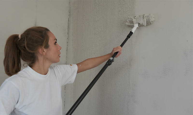 Person i hvit t-skjorte maler lys grå vegg med malerull på forlengerskaft.