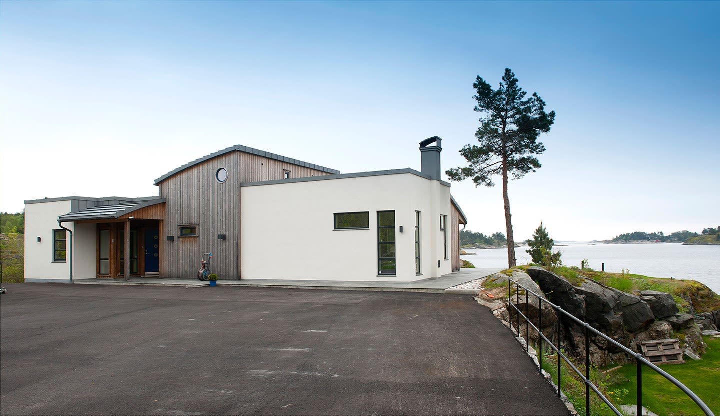 Moderne hvitt hus med trekledning ved klippe- og kystlandskap, grå asfalt, furu og hav